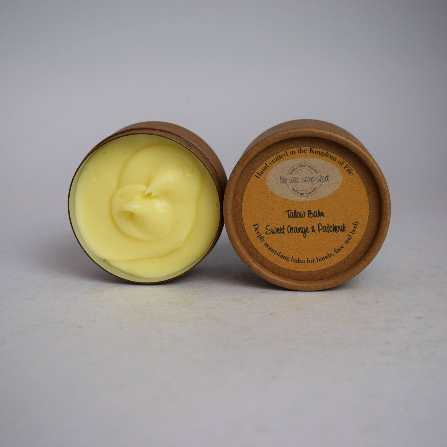Two cardboard containers, one open showing creamy yellow tallow balm on white background